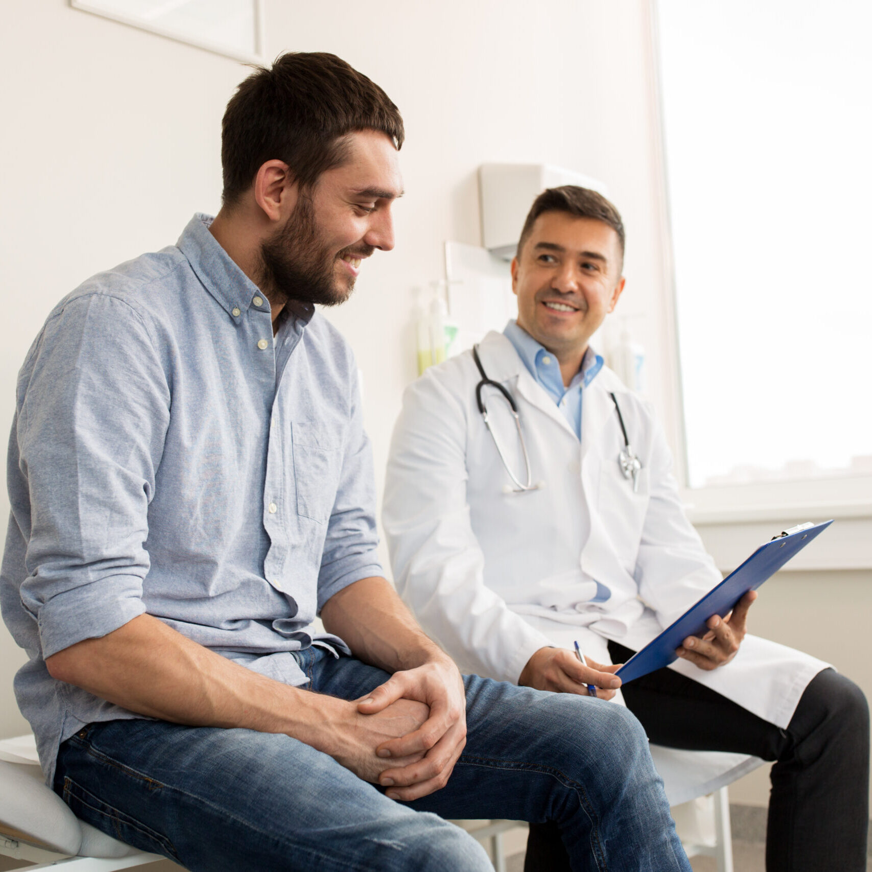 medicine, healthcare and people concept - smiling doctor with clipboard and young man patient meeting at hospital